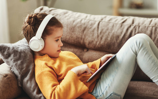 Full body happy girl in casual clothes and headphones s  playing game on tablet while sitting on soft couch at hom - Photo, image