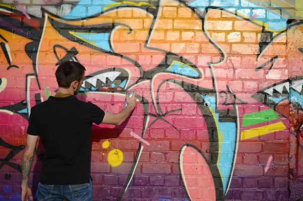 Fiatal graffiti művész hátizsákkal és gázmaszkkal a nyakán színes graffitiket fest rózsaszín tónusokkal a téglafalon. Utcai művészet és kortárs festészet. Szórakozás a fiatalok szubkultúrájában - Fotó, kép