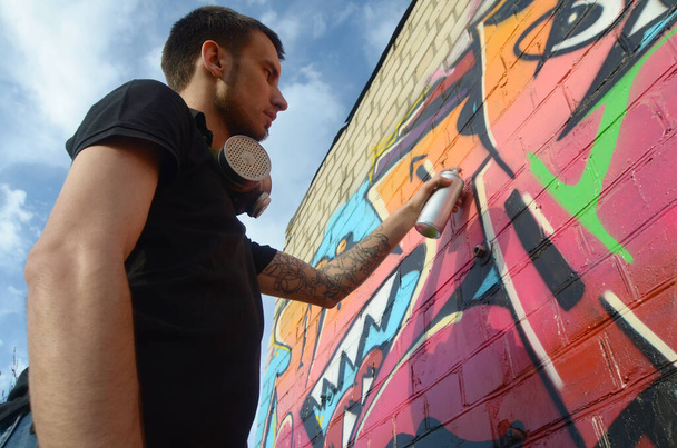 Jovem grafiteiro com mochila e máscara de gás em seu pescoço pinta grafite colorido em tons rosa na parede de tijolo. Arte de rua e processo de pintura contemporânea. Entretenimento na subcultura da juventude
 - Foto, Imagem