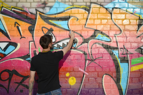 Young graffiti artist with backpack and gas mask on his neck paints colorful graffiti in pink tones on brick wall. Street art and contemporary painting process. Entertainment in youth subculture - Zdjęcie, obraz