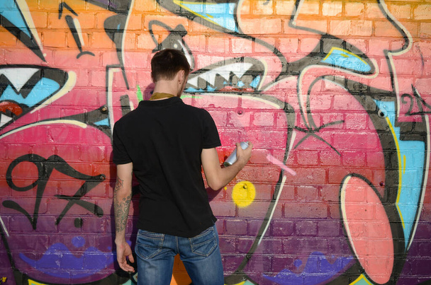 Young graffiti artist with backpack and gas mask on his neck paints colorful graffiti in pink tones on brick wall. Street art and contemporary painting process. Entertainment in youth subculture - Photo, image