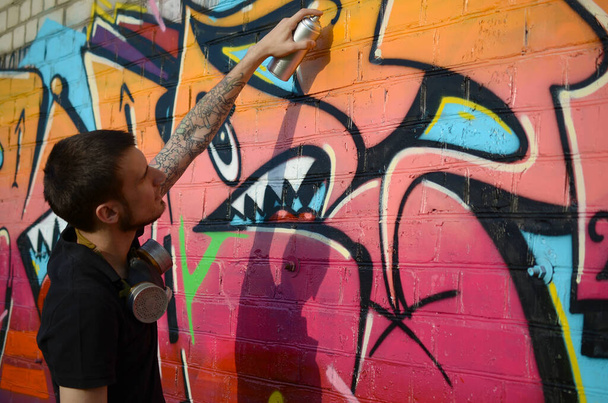 Junger Graffiti-Künstler mit Rucksack und Gasmaske am Hals malt bunte Graffiti in Rosatönen auf Ziegelwände. Street Art und zeitgenössischer Malprozess. Unterhaltung in der Jugend-Subkultur - Foto, Bild