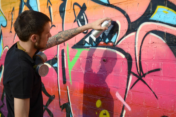 Jonge graffiti kunstenaar met rugzak en gasmasker op zijn nek schildert kleurrijke graffiti in roze tinten op bakstenen muur. Straatkunst en hedendaags schilderproces. Animatie in de jeugdsubcultuur - Foto, afbeelding