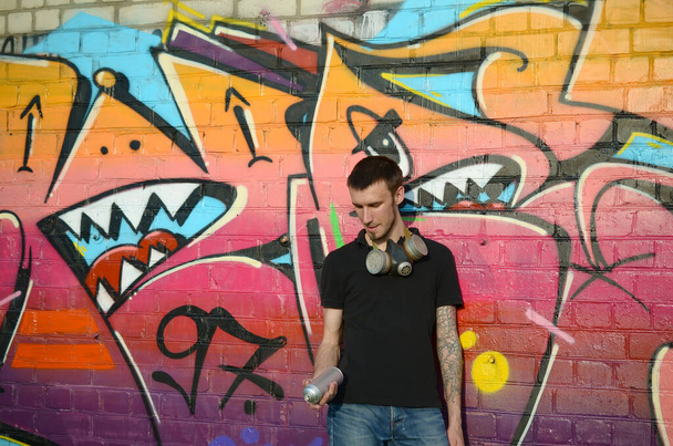 Νεαρός καυκάσιος καλλιτέχνης γκράφιτι σε μαύρο t-shirt με σπρέι αερολύματος κοντά πολύχρωμο ροζ γκράφιτι στον τοίχο από τούβλα. Τέχνη του δρόμου και σύγχρονη ζωγραφική διαδικασία. Ψυχαγωγία στη νεολαία - Φωτογραφία, εικόνα