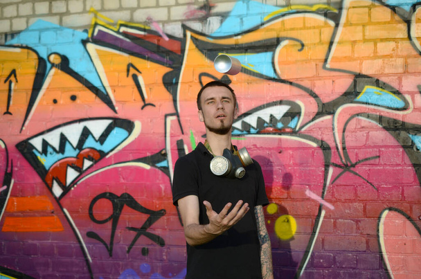Młody graffiti artysta z maską gazową na szyi rzuca puszkę sprayu na kolorowe różowe graffiti na ścianie z cegły. Sztuka uliczna i współczesny proces malowania. Rozrywka w subkulturze młodzieży - Zdjęcie, obraz