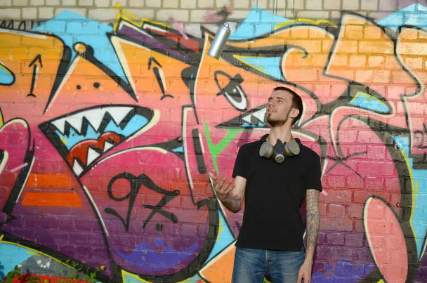 Młody graffiti artysta z maską gazową na szyi rzuca puszkę sprayu na kolorowe różowe graffiti na ścianie z cegły. Sztuka uliczna i współczesny proces malowania. Rozrywka w subkulturze młodzieży - Zdjęcie, obraz