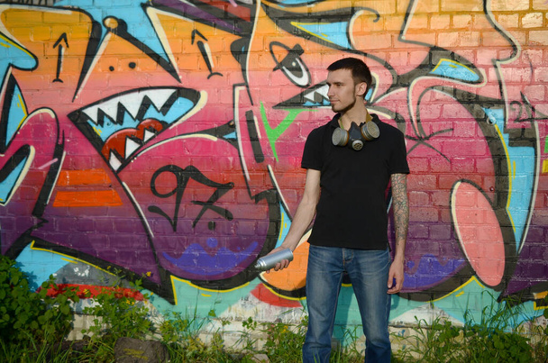 Młody biały artysta graffiti w czarnej koszulce z aerozolową puszką w pobliżu kolorowego różowego graffiti na ceglanej ścianie. Sztuka uliczna i współczesny proces malowania. Rozrywka w subkulturze młodzieży - Zdjęcie, obraz