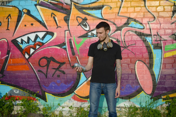 Young caucasian graffiti artist in black t-shirt with aerosol spray can near colorful pink graffiti on brick wall. Street art and contemporary painting process. Entertainment in youth subculture - Photo, Image