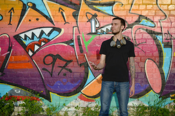 Mladý graffiti umělec s plynovou maskou na krku hodí svou plechovku se sprejem na barevné růžové graffiti na cihlovou zeď. Pouliční umění a současné malířství. Zábava v subkultuře mládeže - Fotografie, Obrázek