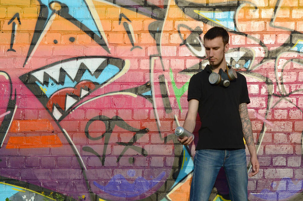 Jonge blanke graffitikunstenaar in zwart t-shirt met spuitbus in de buurt van kleurrijke roze graffiti op bakstenen muur. Straatkunst en hedendaags schilderproces. Animatie in de jeugdsubcultuur - Foto, afbeelding