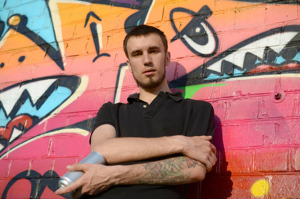Молодой кавказский художник граффити в черной футболке с аэрозольным баллончиком рядом с красочными розовыми граффити на кирпичной стене. Уличное искусство и современный процесс живописи. Развлечения в молодежной субкультуре
 - Фото, изображение