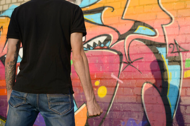 Rückansicht eines jungen kaukasischen Mannes mit Messingknöchelchen an der Hand gegen eine Mauer aus Ghettoziegeln mit Graffiti-Bildern. Konzept krimineller Kräfte und Aggressionsaufladung. Klassische Gangster-Nahkampfwaffe - Foto, Bild