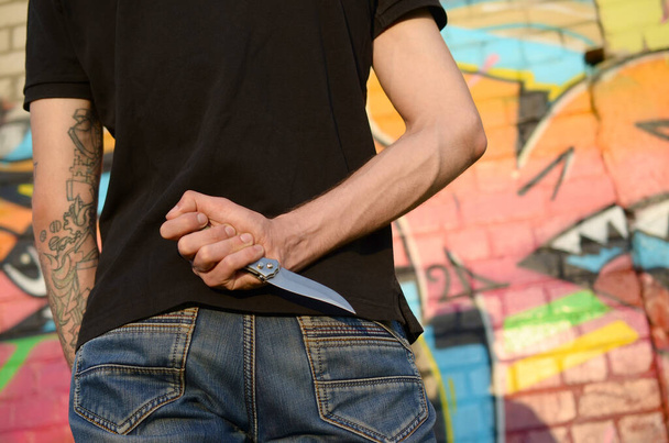 Rückansicht eines jungen kaukasischen Mannes mit einem Messer in der Hand gegen eine Mauer aus Ghettoziegeln mit Graffiti-Bildern. Konzept krimineller Kräfte und Aggressionsaufladung. Klassische Killer-Nahkampfwaffe - Foto, Bild