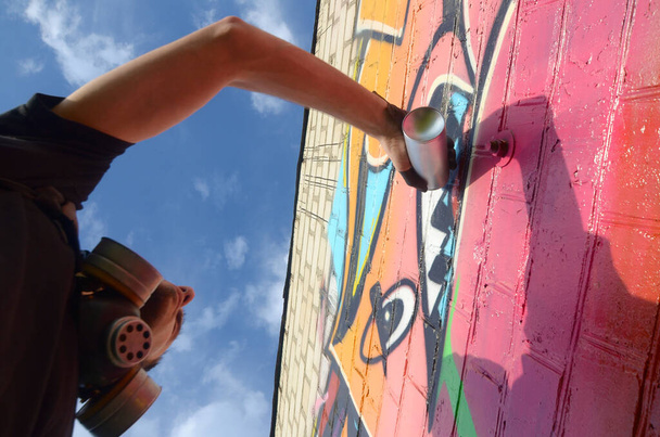 Młody graficiarz z plecakiem i maską gazową na szyi maluje kolorowe graffiti w różowych odcieniach na ceglanej ścianie. Sztuka uliczna i współczesny proces malowania. Rozrywka w subkulturze młodzieży - Zdjęcie, obraz