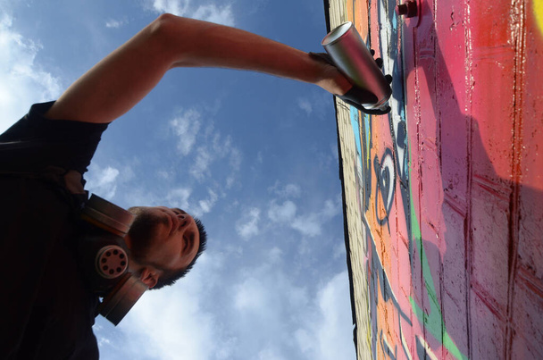 Jonge graffiti kunstenaar met rugzak en gasmasker op zijn nek schildert kleurrijke graffiti in roze tinten op bakstenen muur. Straatkunst en hedendaags schilderproces. Animatie in de jeugdsubcultuur - Foto, afbeelding
