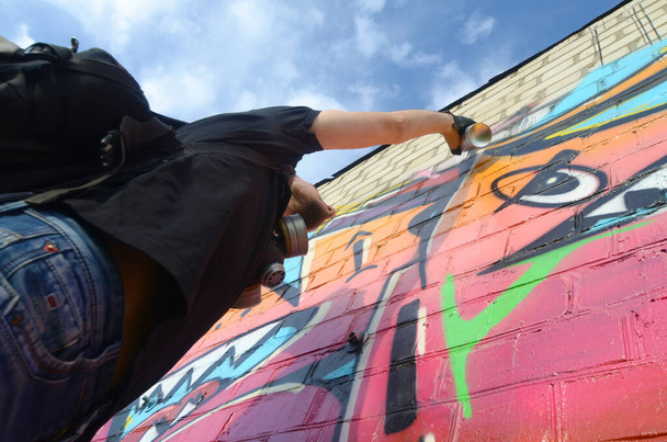 Молодий художник графіті з рюкзаком і газовою маскою на шиї малює барвисті графіті в рожевих тонах на цегляній стіні. Вуличне мистецтво та процес сучасного живопису. Розваги в молодіжній субкультурі
 - Фото, зображення