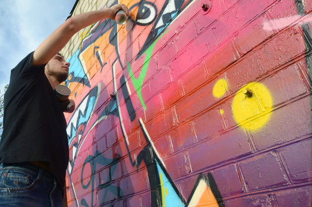 Nuori graffititaiteilija, jolla on reppu ja kaasunaamari kaulassaan, maalaa värikkäitä graffiteja vaaleanpunaisilla sävyillä tiiliseinään. Katutaide ja nykyaikainen maalausprosessi. Viihde nuorisoalakulttuurissa
 - Valokuva, kuva
