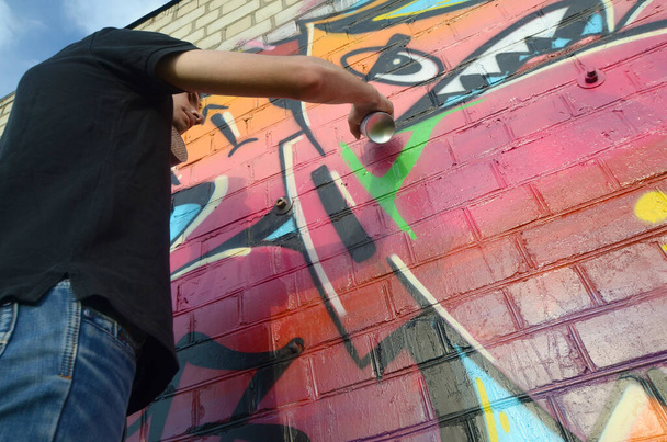 Молодой граффити-художник с рюкзаком и противогазом на шее рисует красочные граффити в розовых тонах на кирпичной стене. Уличное искусство и современный процесс живописи. Развлечения в молодежной субкультуре
 - Фото, изображение