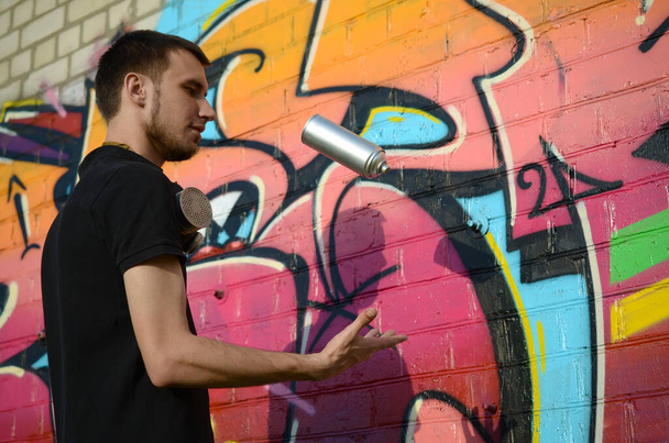 Nuori graffiti taiteilija kaasunaamari kaulassaan heittää spray tölkki vastaan värikäs vaaleanpunainen graffiti tiiliseinällä. Katutaide ja nykyaikainen maalausprosessi. Viihde nuorisoalakulttuurissa
 - Valokuva, kuva