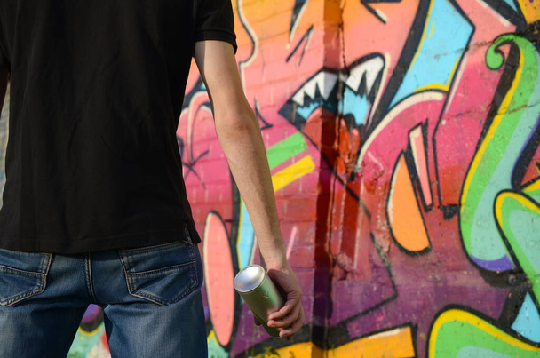 Νεαρός καλλιτέχνης γκράφιτι με σακίδιο και μάσκα αερίου στο λαιμό ζωγραφίζει πολύχρωμα γκράφιτι σε ροζ τόνους στον τοίχο από τούβλα. Τέχνη του δρόμου και σύγχρονη ζωγραφική διαδικασία. Ψυχαγωγία στη νεολαία - Φωτογραφία, εικόνα