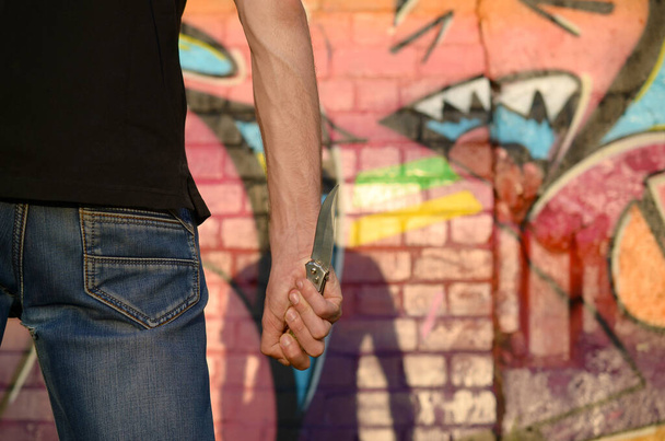 Achteraanzicht van de jonge blanke man met mes in zijn hand tegen getto bakstenen muur met graffiti schilderijen. Concept van criminele krachten en agressie aanklacht. Klassieke moordenaars melee wapen - Foto, afbeelding