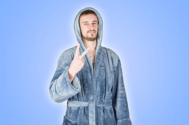 Portrait of young caucasian bearded man in blue bathrobe shows victory or peace gesture isolated on blue background. Happy emotion - Photo, image