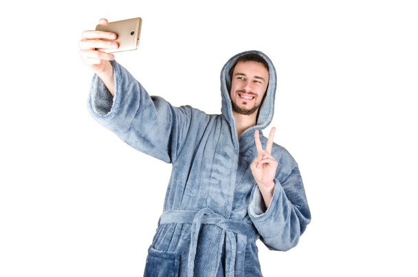 Portrait of young caucasian bearded man in blue bathrobe shows victory gesture and makes selfie isolated on white background. Happy emotion concept - Фото, изображение