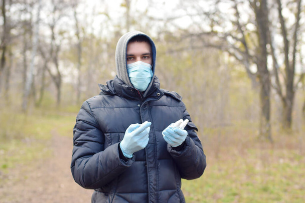Young man in protective mask shows sanitizer spray bottles outdoors in spring wood. Concept of usage liquid anticeptic spray during quarantine - Φωτογραφία, εικόνα