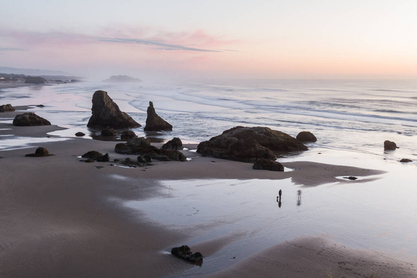 os pinks macios que refletem na areia molhada enquanto as ondas se movem para trás e para a frente com as rochas grandes que estão altas adicionando o drama a esta cena litoral em Bandon, Oregon
 - Foto, Imagem