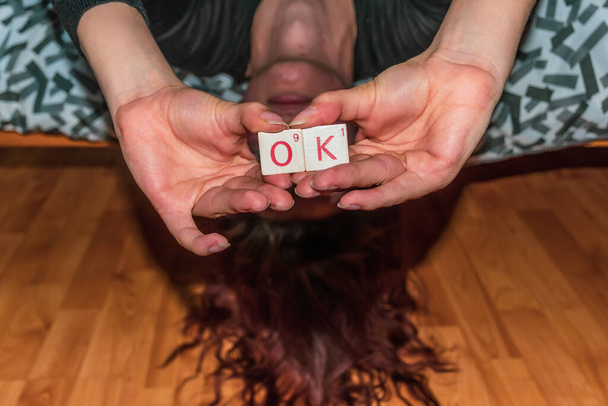 Um close-up de uma jovem mulher ruiva caucasiana irreconhecível deitada de cabeça para baixo em uma cama segurando as telhas de madeira dizendo "ok" na frente de seu rosto
 - Foto, Imagem