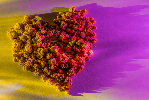 Eine Nahaufnahme von Getreidekörnern in Herzform auf einem Papierblatt, das von den gegenüberliegenden Seiten mit leuchtendem Magenta und gelbem Licht beleuchtet wird - Foto, Bild