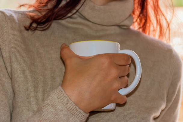A close-up shot of an unrecognizable young Caucasian redhead woman wearing a beige sweater holding a cup of warm coffee with one hand close to her chest - Photo, Image