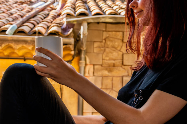 A close-up shot of an unrecognizable young Caucasian redhead woman sitting on a windowsill holding a cup of hot beverage and smiling with an old French building in the background - Photo, Image