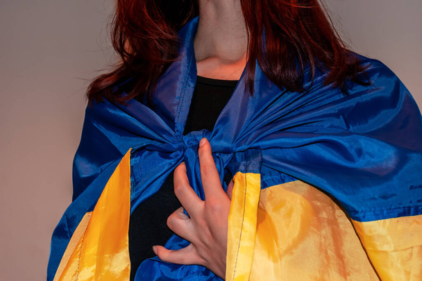 A close-up shot of an unrecognizable young Caucasian redhead woman wearing a Ukrainian national flag as a cape with her hand on the knot - Foto, Bild