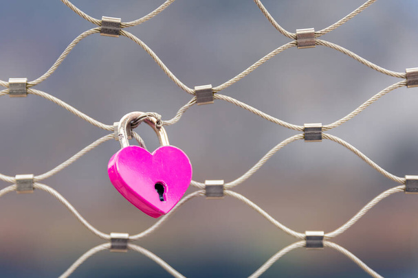 Close focus on pink lock in heart shape hanging on metal net fence with blurry background for loving representation. - Zdjęcie, obraz