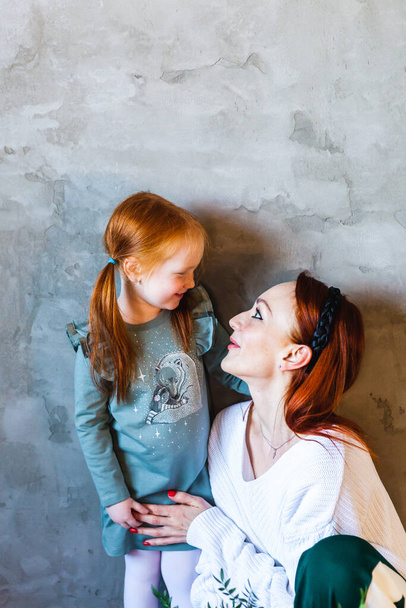 Little girl with red hair, braided ponytails, in a dress, stands near the wall and hugs mom, communicating with a parent, relationships, family, attention, emotions, feelings, significance, upbringing - Foto, Bild