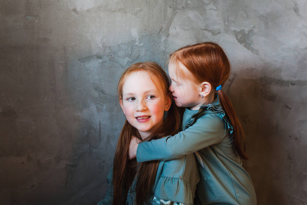 A little girl with red hair hugs her sister, whispers a secret in her ear, plays, laughs, family relations, growing up, games, mom and dad, parents, home - Zdjęcie, obraz
