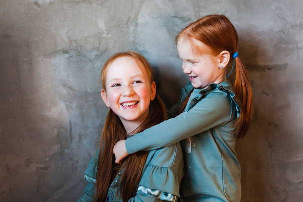A little girl with red hair hugs her sister, whispers a secret in her ear, plays, laughs, family relations, growing up, games, mom and dad, parents, home - Фото, изображение