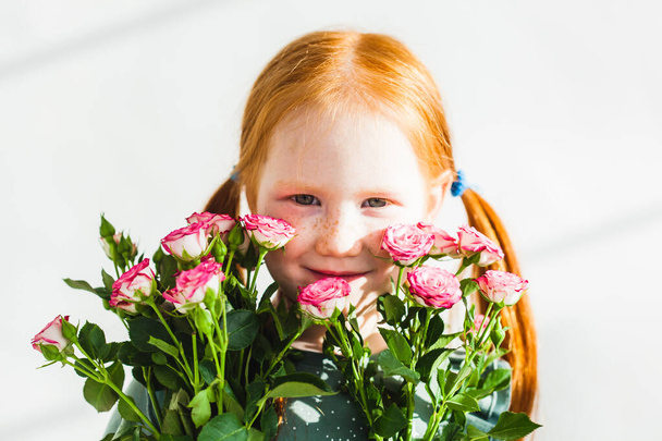 Little girl with red hair, braided ponytails, in a dress, stands near the wall and holds green plants, and roses, pistachio, emotions, childhood, parents, mother, interior, joy - Photo, image