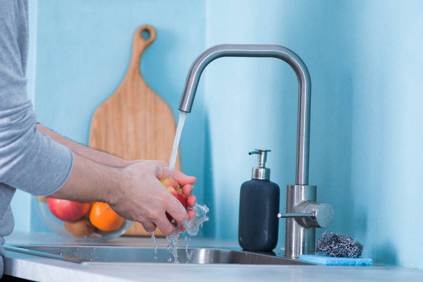 A man washes fruit in a sink in a kitchen with blue walls. Against the background of fruits. Men's hands wash an apple - Фото, зображення