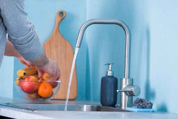 A man washes fruit in a sink in a kitchen with blue walls. Against the background of fruits. Men's hands wash an apple - Фото, зображення