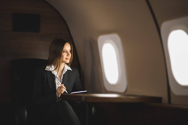 Šťastná hnědovlasá mladá běloška, držící tablet, sedící na sedadle v soukromém tryskáči, dívající se na okno letadla a usmívající se. - Fotografie, Obrázek