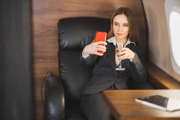 A confident successful caucasian business lady sitting on a private plane with a glass of champagne and takes a selfie on her phone. Business, success concept. - Фото, изображение