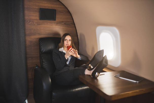 Serious young businesswoman sitting in first class of private plane with her feet on table, holding mobile phone, looking to side with concerned worried expression. - Fotografie, Obrázek