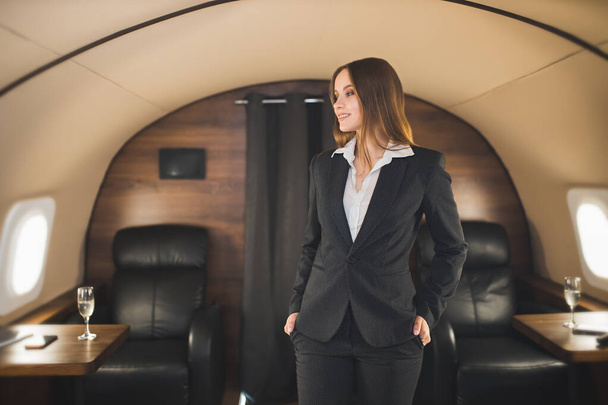 Portrait of attractive happy young brown haired business woman with charming smile, wearing suit standing in comfortable first class cabin. Beautiful lady in private jet. Success, business concept. - Photo, image