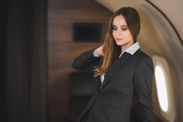 Beautiful caucasian young brown haired lady with nice makeup, wearing stylish black suit, looks down, touches her hair and gently smiles, poses in private aircraft. Traveling at first class. - Foto, Imagem