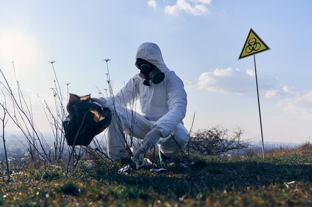Male research ecologist in protective suit and gas mask picking up garbage, putting into garbage bag in field with biohazard sign. Concept of ecology, environmental pollution and biological hazard. - Photo, Image