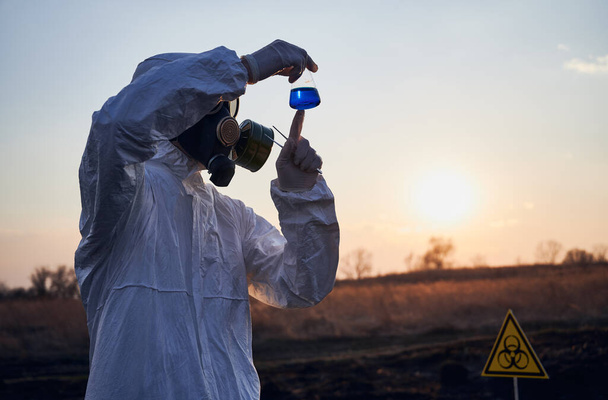 Мужчина-эколог в защитном костюме, противогазе и перчатках держит пробирку с голубой жидкостью во время изучения образцов почвы на выжженной территории с биологическим знаком опасности. Концепция экологии и исследований
 - Фото, изображение