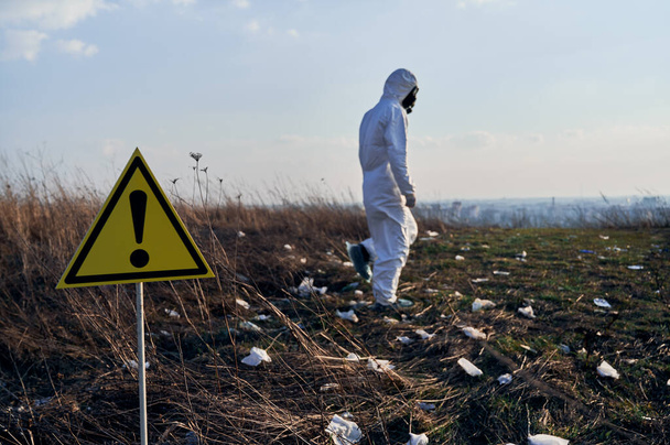 Yellow triangle with black exclamation mark warning about danger and hazards in field with trash. Warning sign with male ecologist in radiation suit on blurred background. Concept of ecology - Photo, Image