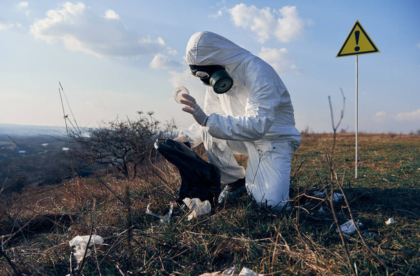 Widok z boku na ekologa noszącego biały kombinezon ochronny, maskę gazową, kucanie zbiera śmieci do czarnego worka na śmieci na zewnątrz w słoneczny dzień. Znak ostrożności za nim. Pojęcie ekologii, zanieczyszczenie - Zdjęcie, obraz